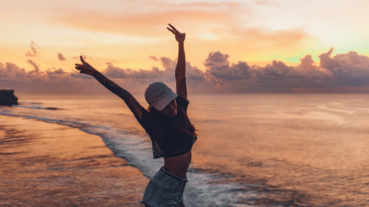 Side view fit lady in casual shorts and cap enjoying evening on seashore while standing with arms raised against calm endless sea and majestic bright cloudy orange sky during sunset